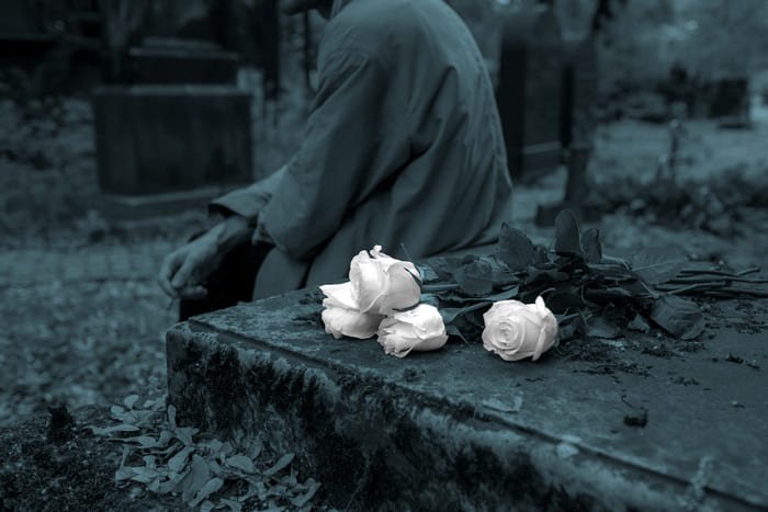 Eine Person sitzt vor einem Grabstein, auf dem weiße Rosen liegen – Symbol für die Bestattungsvorsorge von RIANEVA, die finanzielle Sicherheit und Entlastung im Trauerfall bietet.